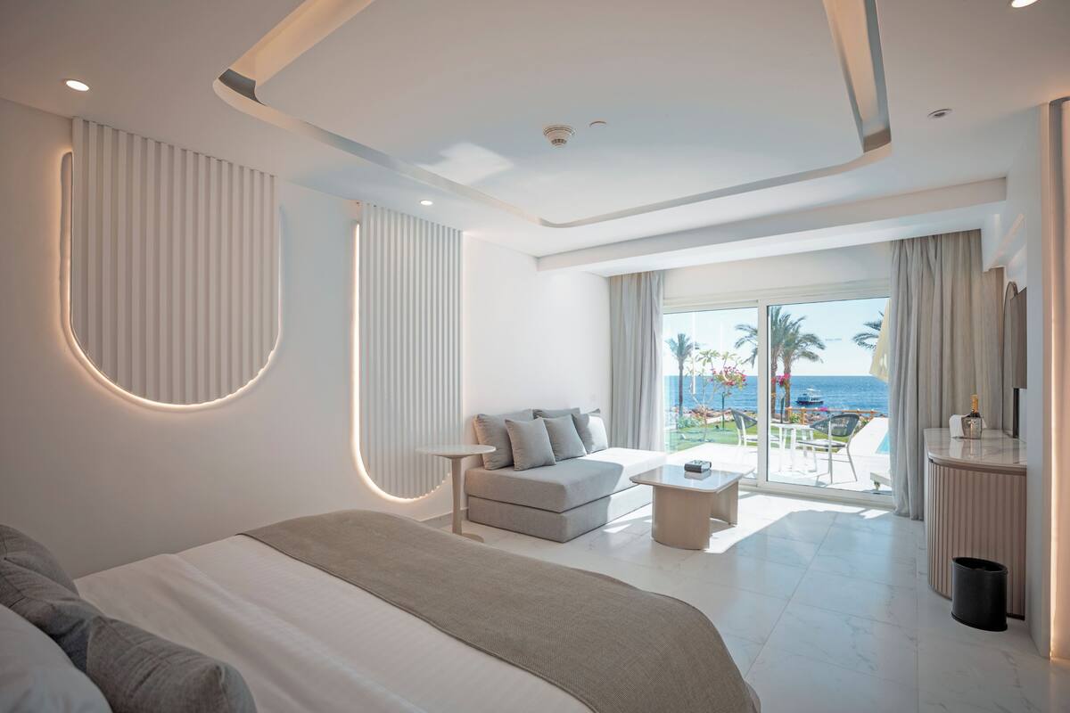A modern hotel room with a large bed, a sofa, a small table, and a balcony with a view of the sea.