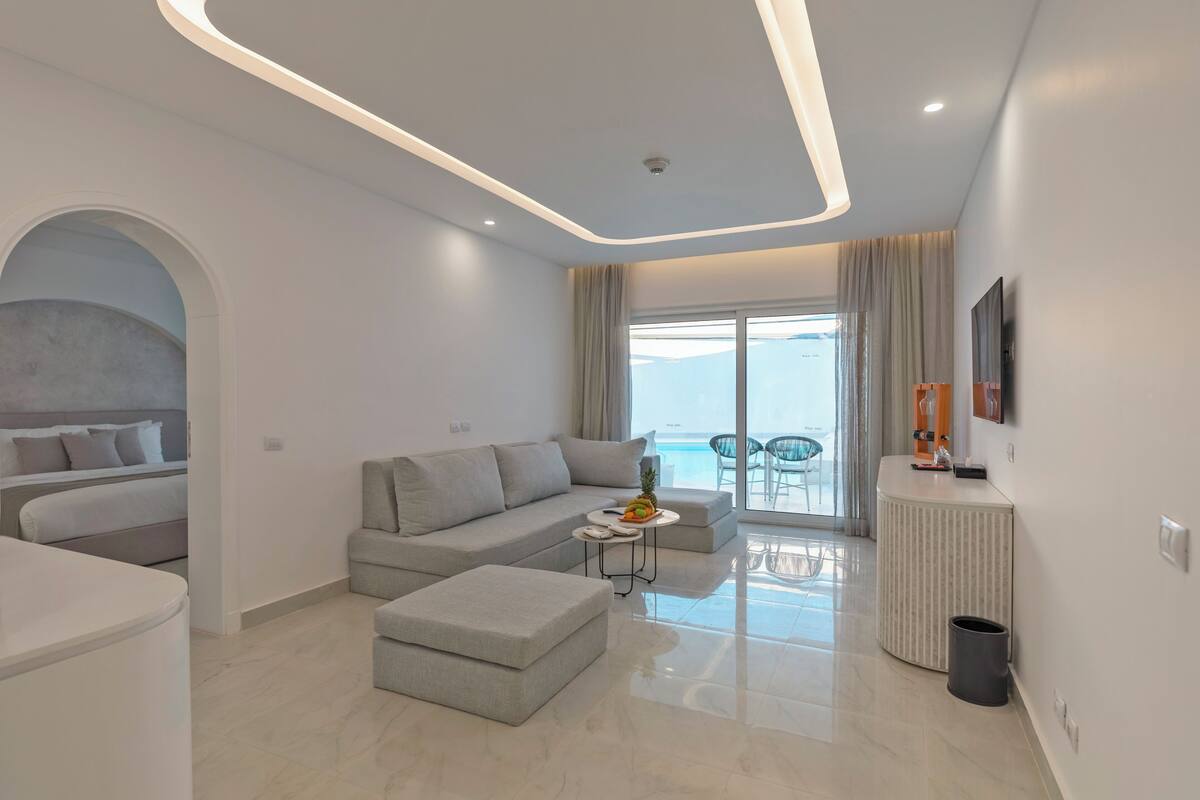 A modern living room with a grey sofa, a small round table with fruit, and a view of the sea through large windows.