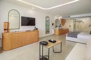 A modern hotel room with a wooden reception desk, a large mirror, a flat-screen TV, a bed with a gray blanket, and a glass shower enclosure.