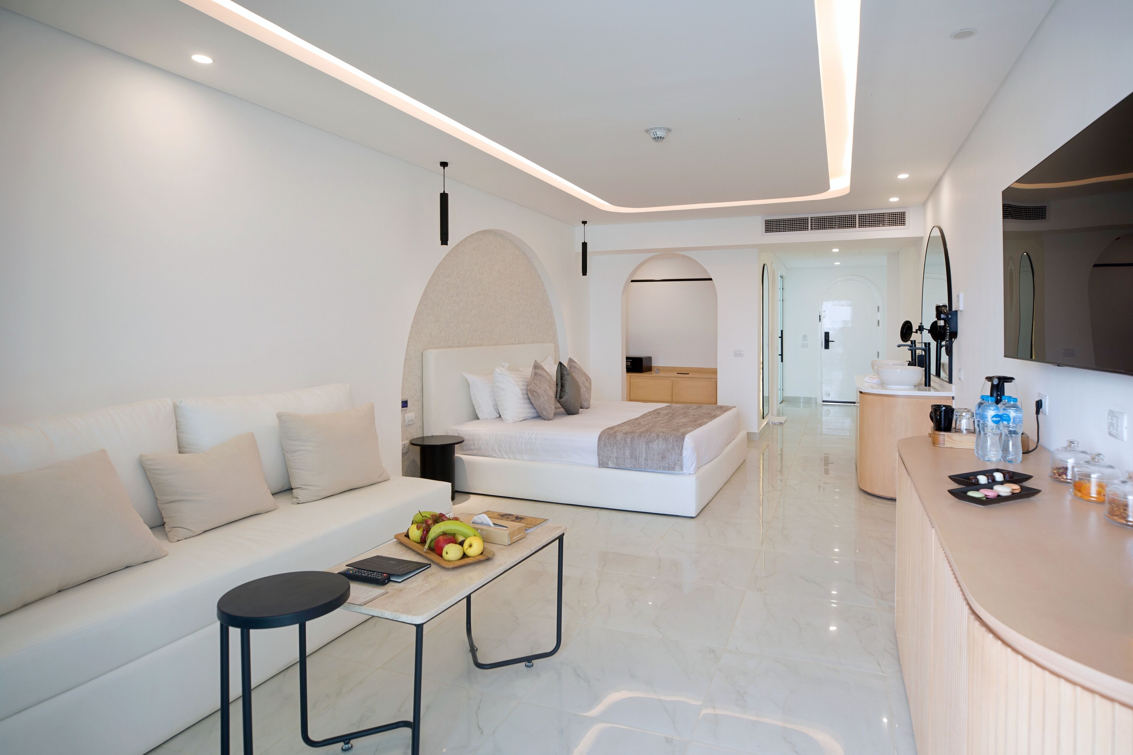 A modern hotel room with a white sofa, a bed with a wooden headboard, a small table with fruits, a bathroom with a mirror, and a reception desk.