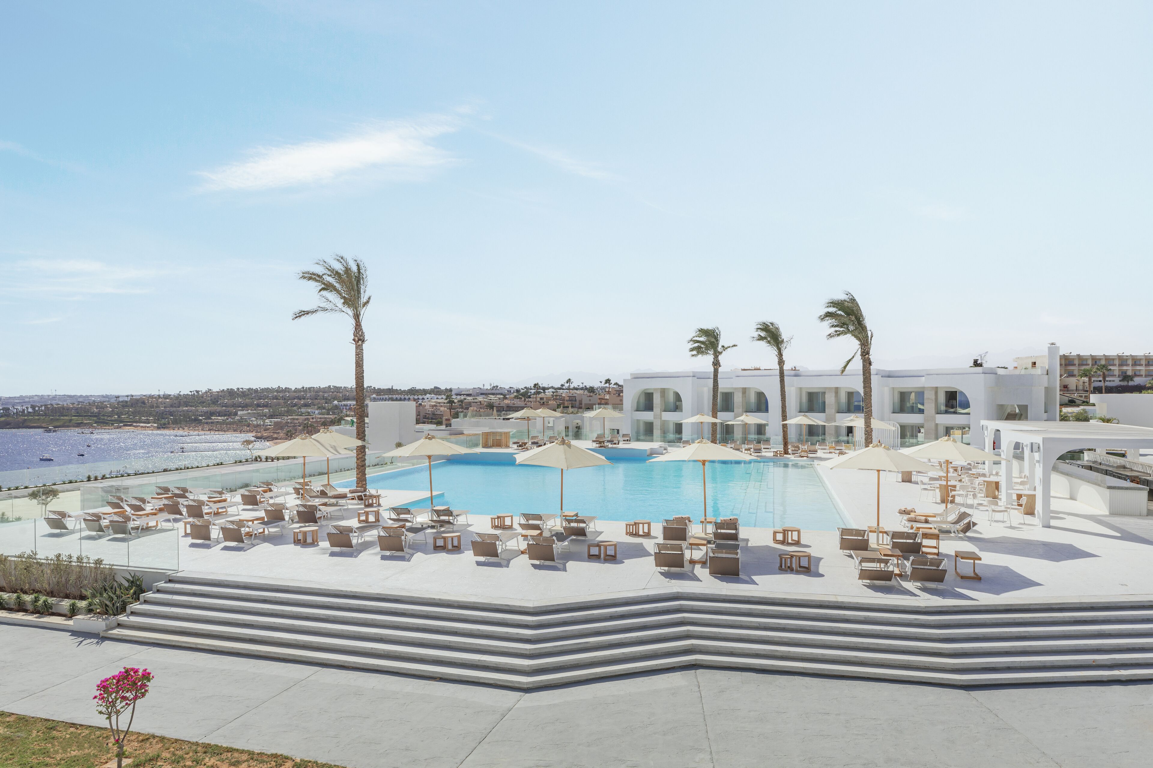 A pool area with lounge chairs and umbrellas, a clear view of the sea, and a modern building in the background.