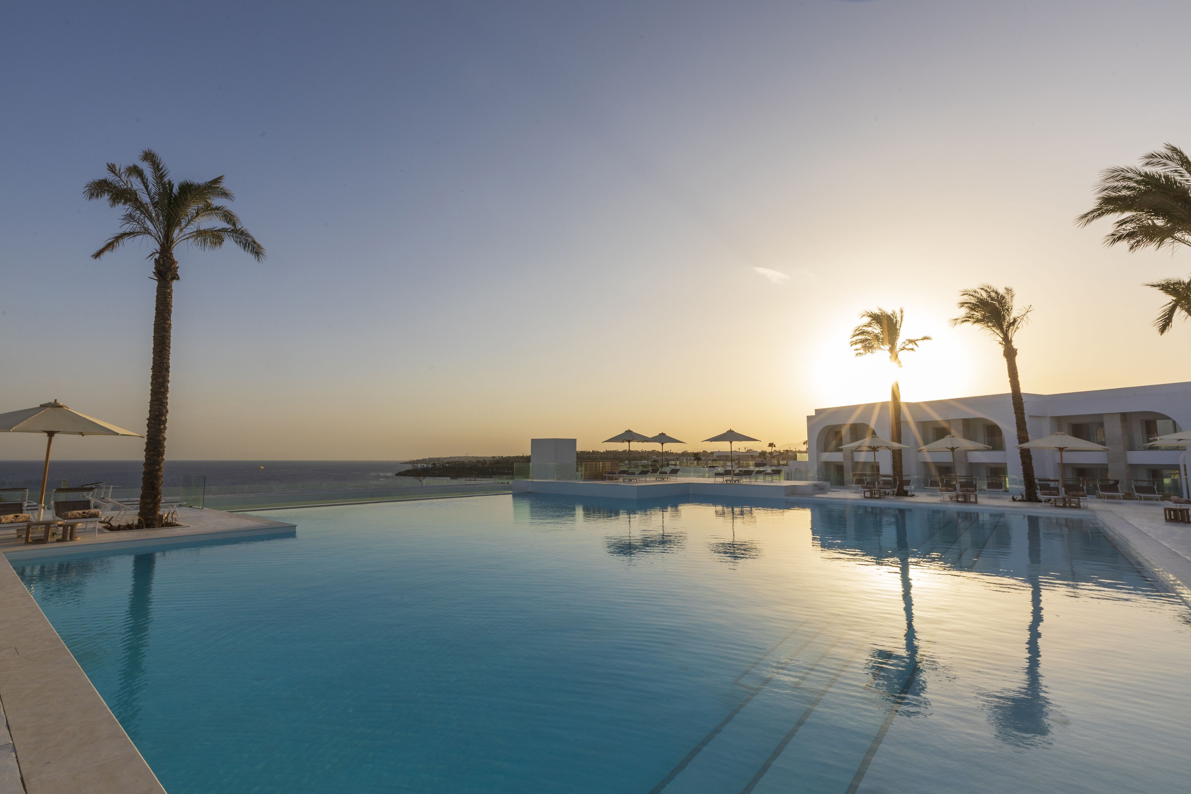 A swimming pool with palm trees and umbrellas, overlooking the ocean at sunset.