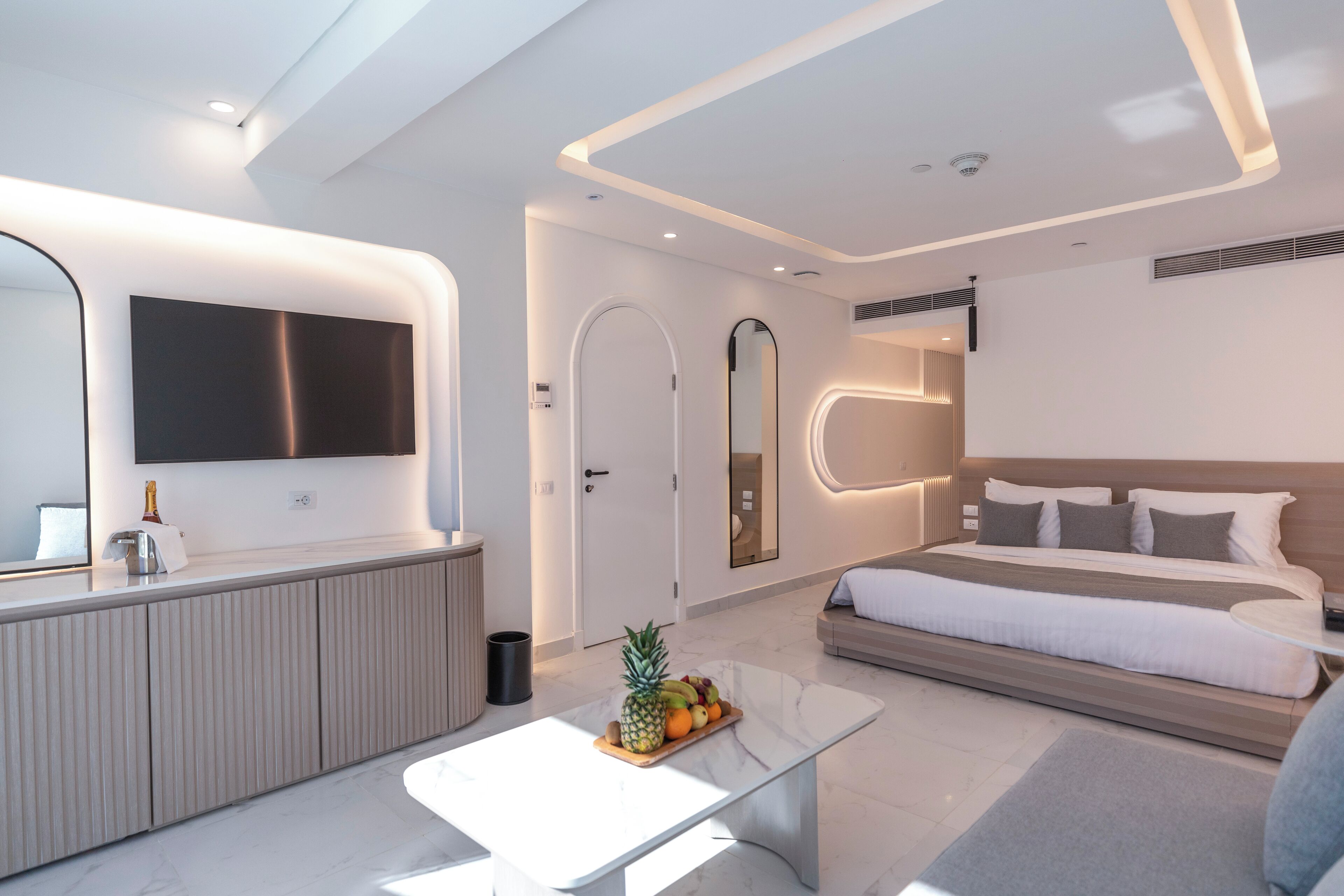 A modern hotel room with a large bed, a bedside table with fruit, a mirror, and a TV above the bed.