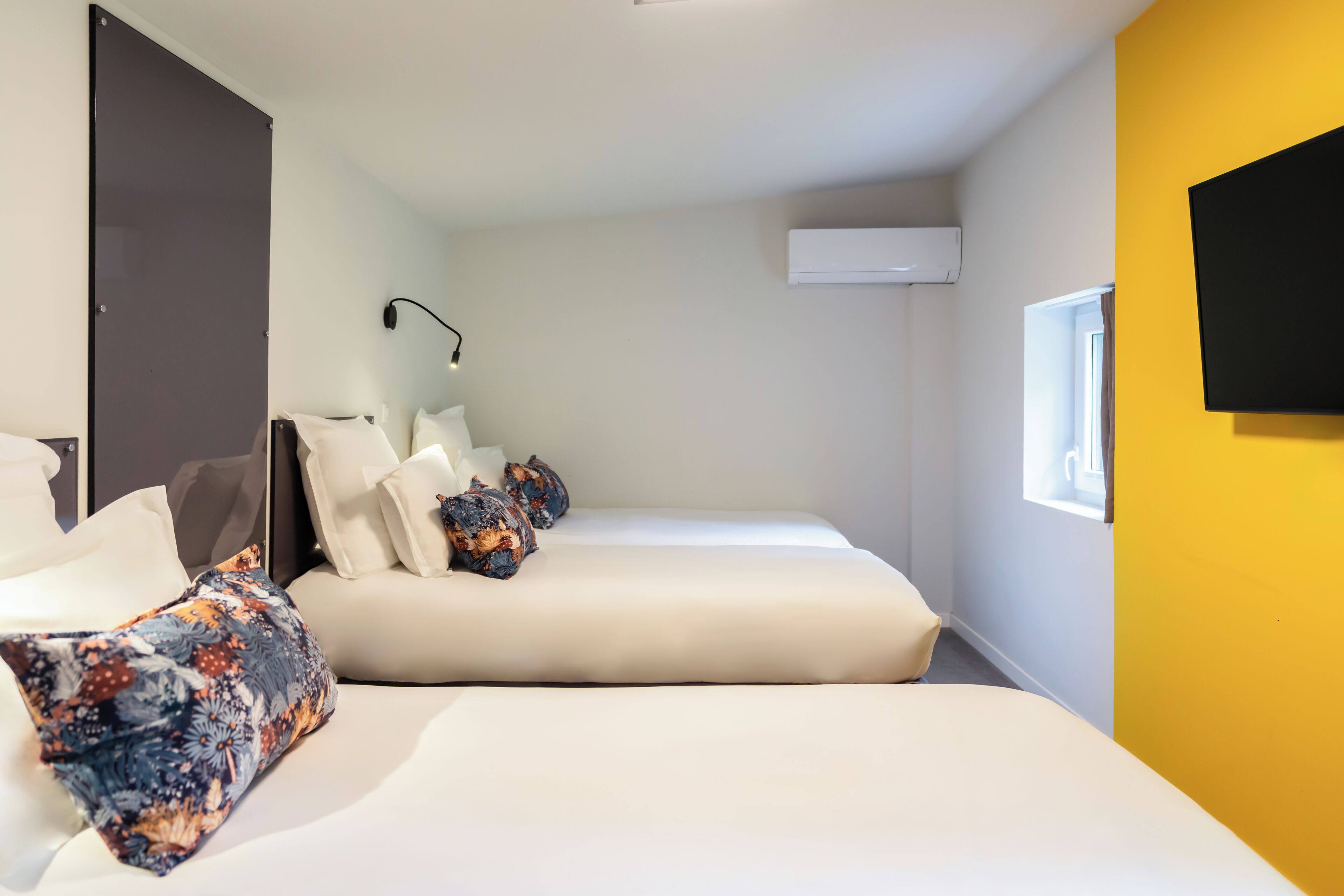 A hotel room with two beds, a flat-screen TV, an air conditioning unit, and a window with curtains.