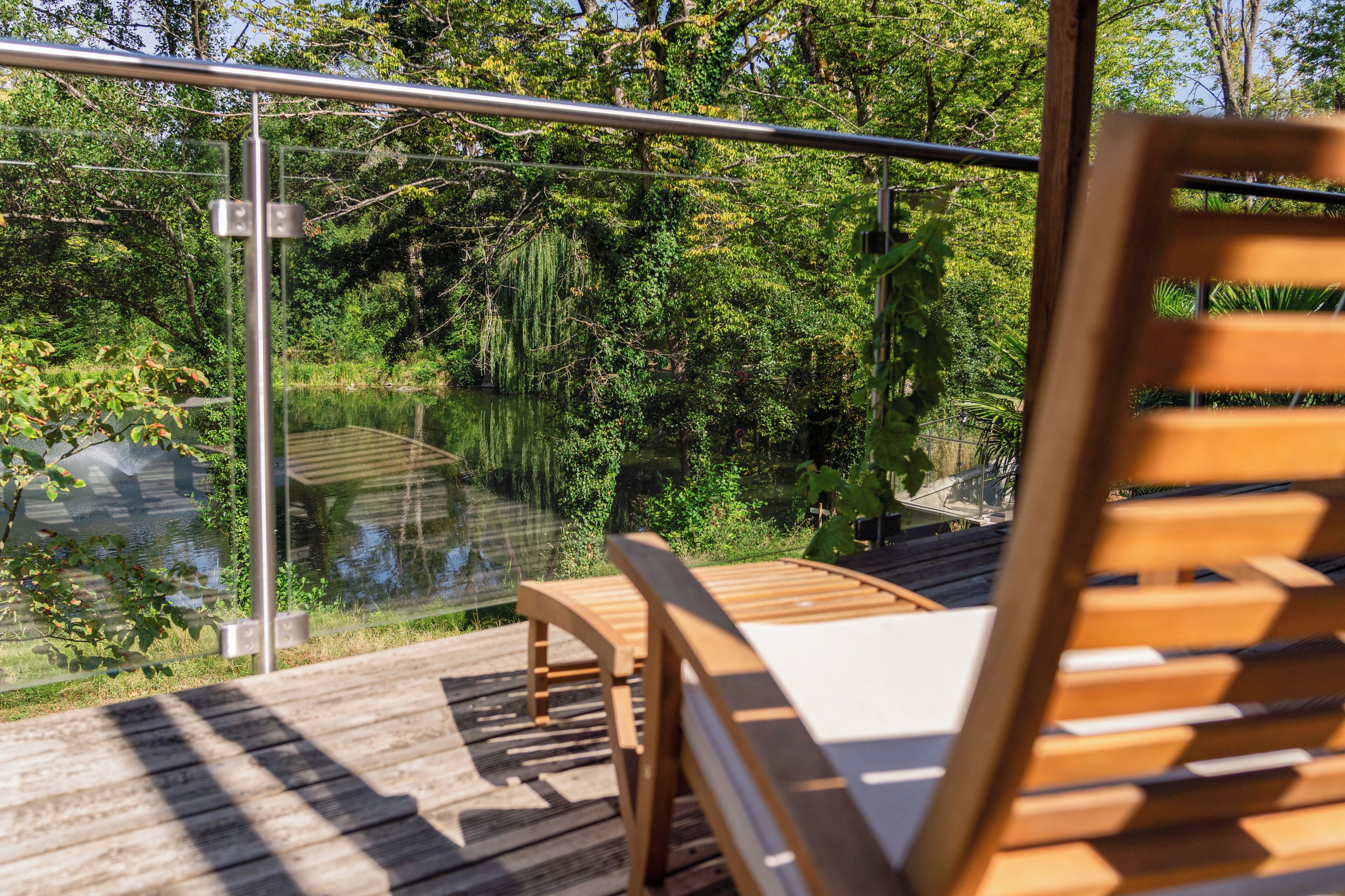 A wooden deck with a glass railing overlooking a pond and lush greenery.