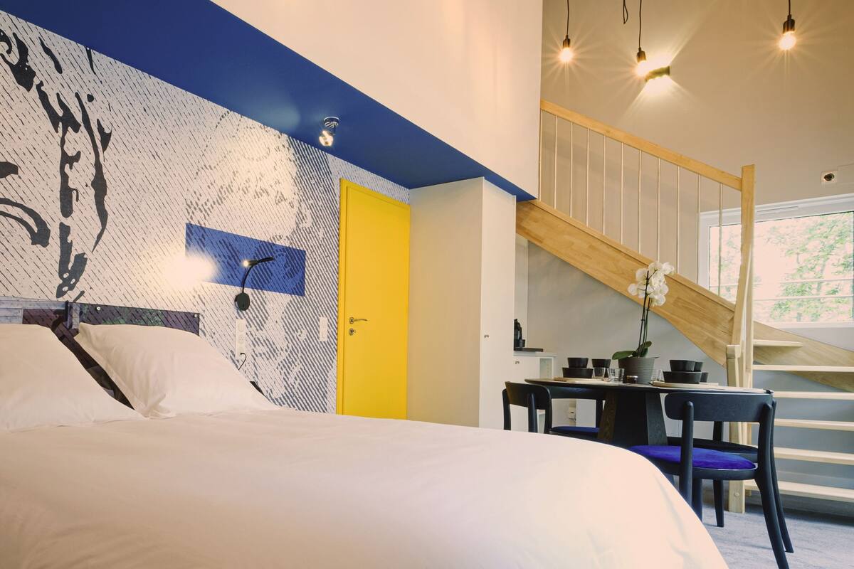 Un dormitorio con cama, una puerta amarilla, una escalera y una zona de comedor con sillas y una mesa.