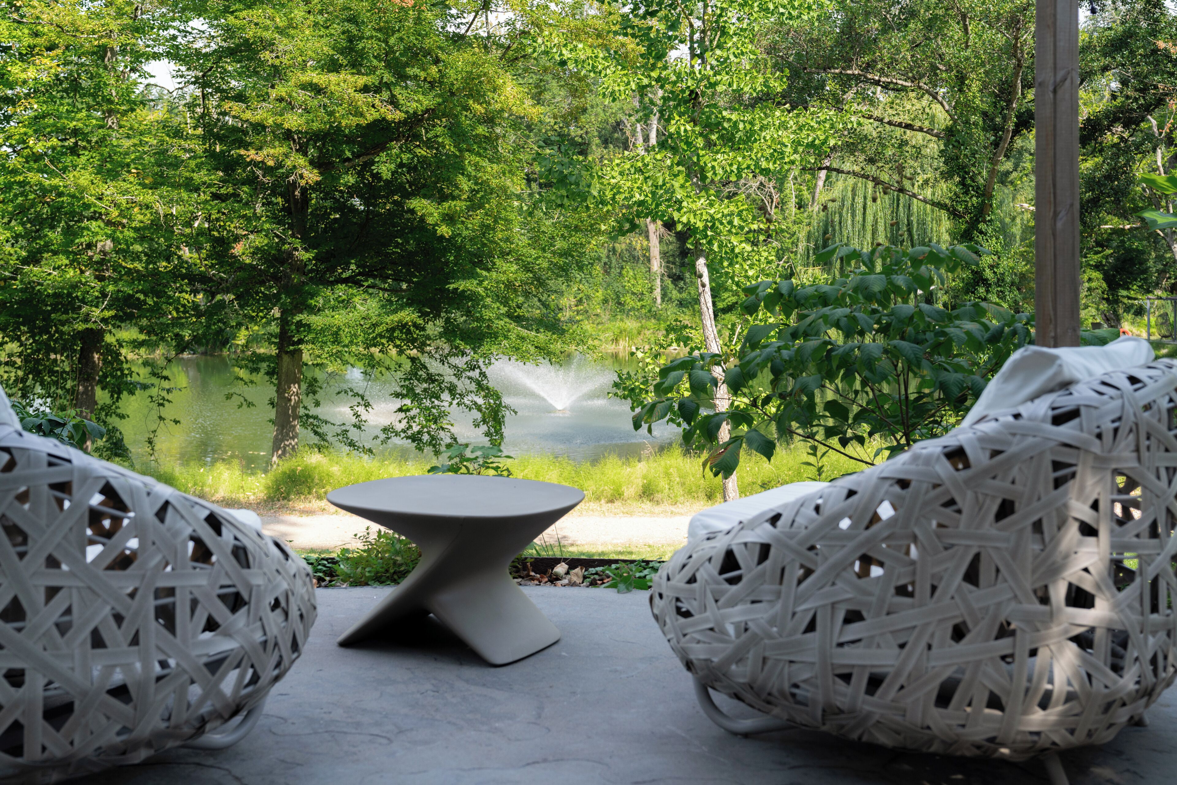 Uma área de estar tranquila ao ar livre, com uma mesa e duas cadeiras de vime, com vista para um lago rodeado por vegetação exuberante.