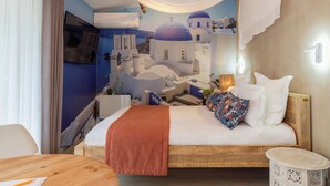 Hotellihuoneessa on sänky, puinen pöytä, pieni pyöreä pöytä ja seinämaalaus, jossa on kuvattuna rannikkokylä.