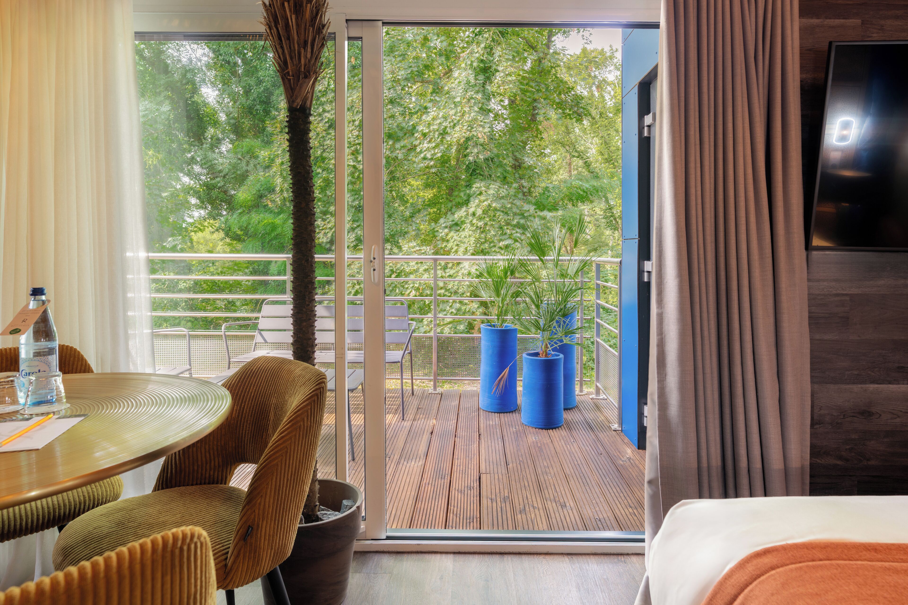 A hotel room with a balcony, a dining table with chairs, a potted plant, and a TV mounted on the wall.