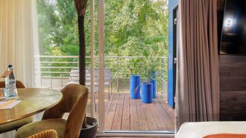 A hotel room with a balcony, a dining table with chairs, a potted plant, and a TV mounted on the wall.