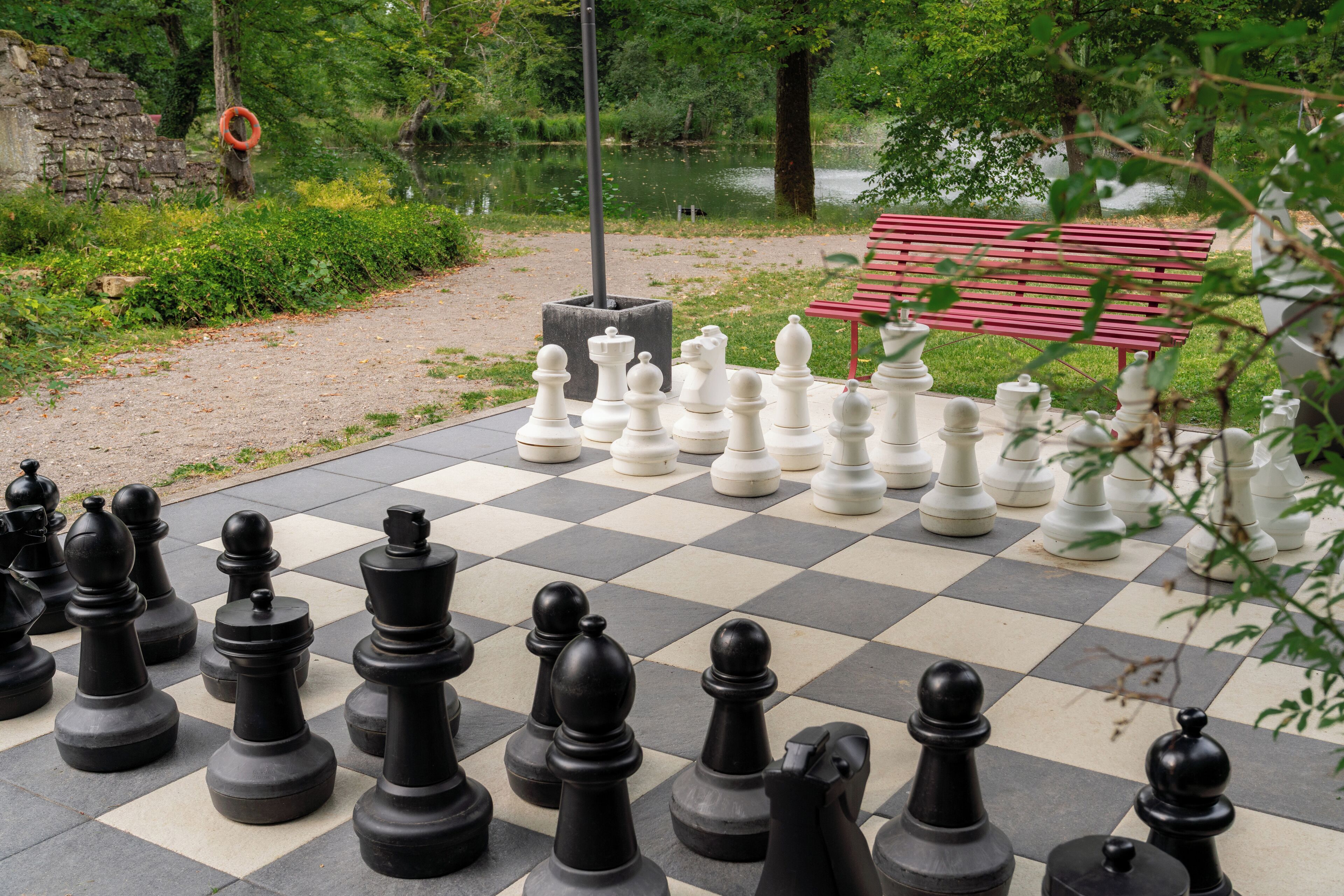 A large outdoor chessboard set up in a park with a bench in the background.