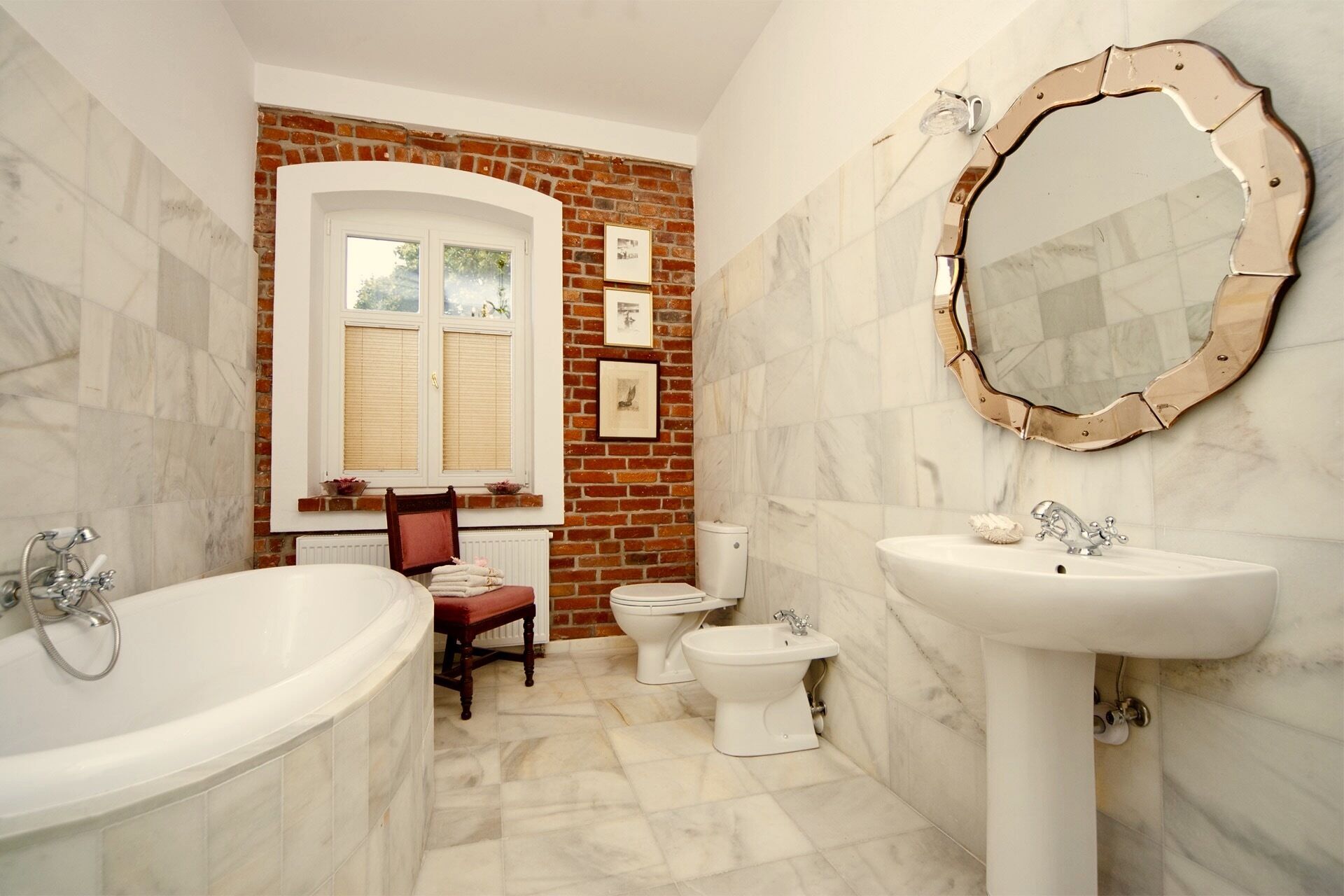 Ein Badezimmer mit Badewanne, Waschbecken, Toilette und Bidet. Es gibt ein Fenster mit Jalousien, einen Sessel und einen Spiegel mit dekorativem Rahmen.