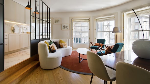 A modern living room with a white sofa, a round coffee table, and a dining table with chairs. There are large windows with curtains, a kitchen area with marble countertops, and pendant lighting. - Amazing 2 Bedroom Apt w/ Great Views, 2 min Walk From Douro River in City Center (Porto)