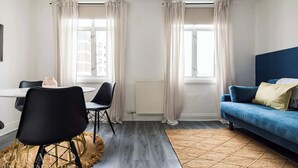 A modern living room with a blue sofa, a dining table with chairs, and large windows with curtains.