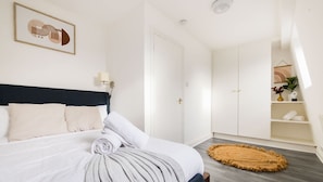 A bedroom with a bed, a headboard, a bedside lamp, a door, a wardrobe, a shelf with a plant, and a round rug.