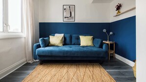 A room with a blue sofa, a woven rug, a framed picture on the wall, and a small side table with a lamp.