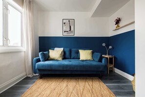 A room with a blue sofa, a woven rug, a framed picture on the wall, and a small side table with a lamp.