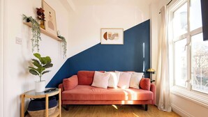 A room with a blue accent wall, a pink sofa, a small side table with a plant, and a window with curtains.