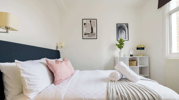 A neatly made bed with white and pink pillows, a grey throw blanket, and a rolled towel. There are two framed artworks on the wall, a potted plant, and a white shelf with a lamp and a basket. - 1 Bedroom Flat - Shepherds Bush (Greater London)