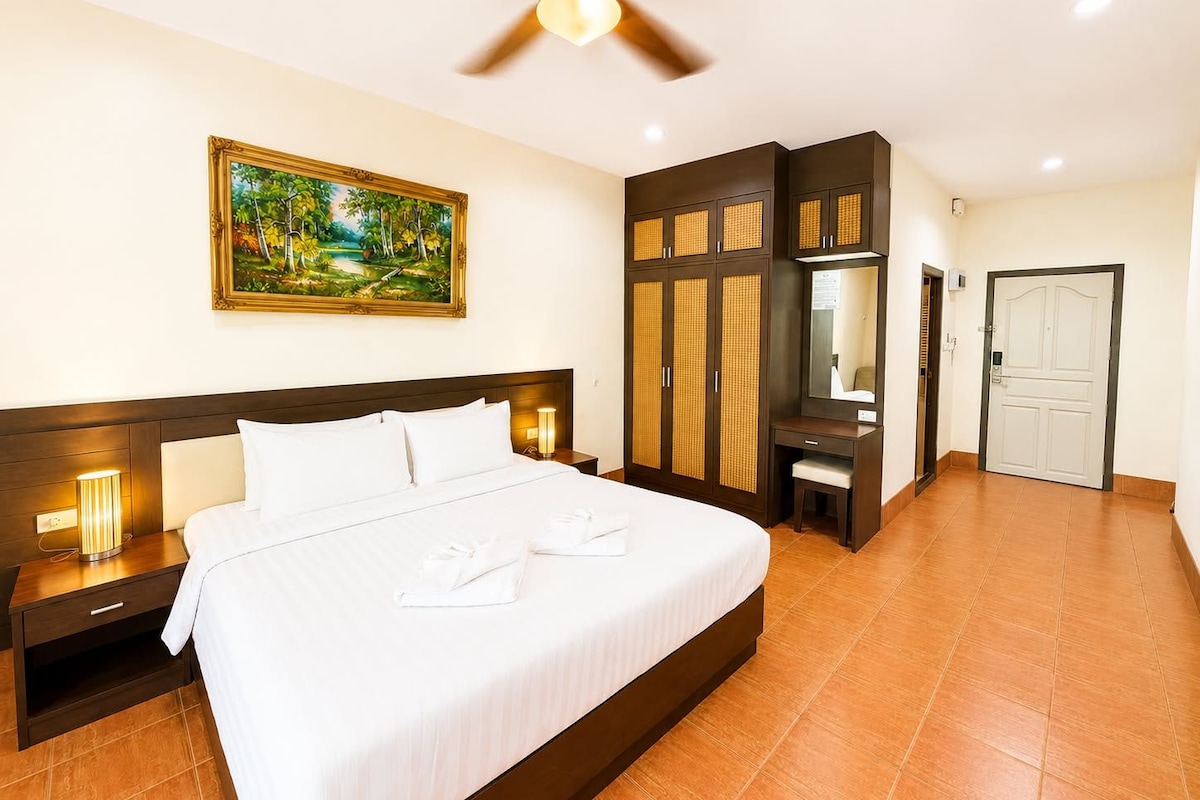 A hotel room with a bed, a desk, a wardrobe, a ceiling fan, and a painting on the wall.