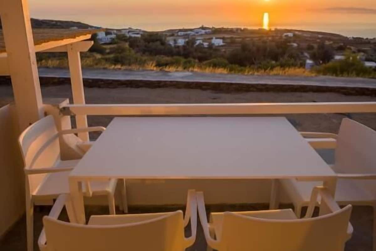 A terrace with a table and chairs, overlooking a sunset over a landscape with buildings.