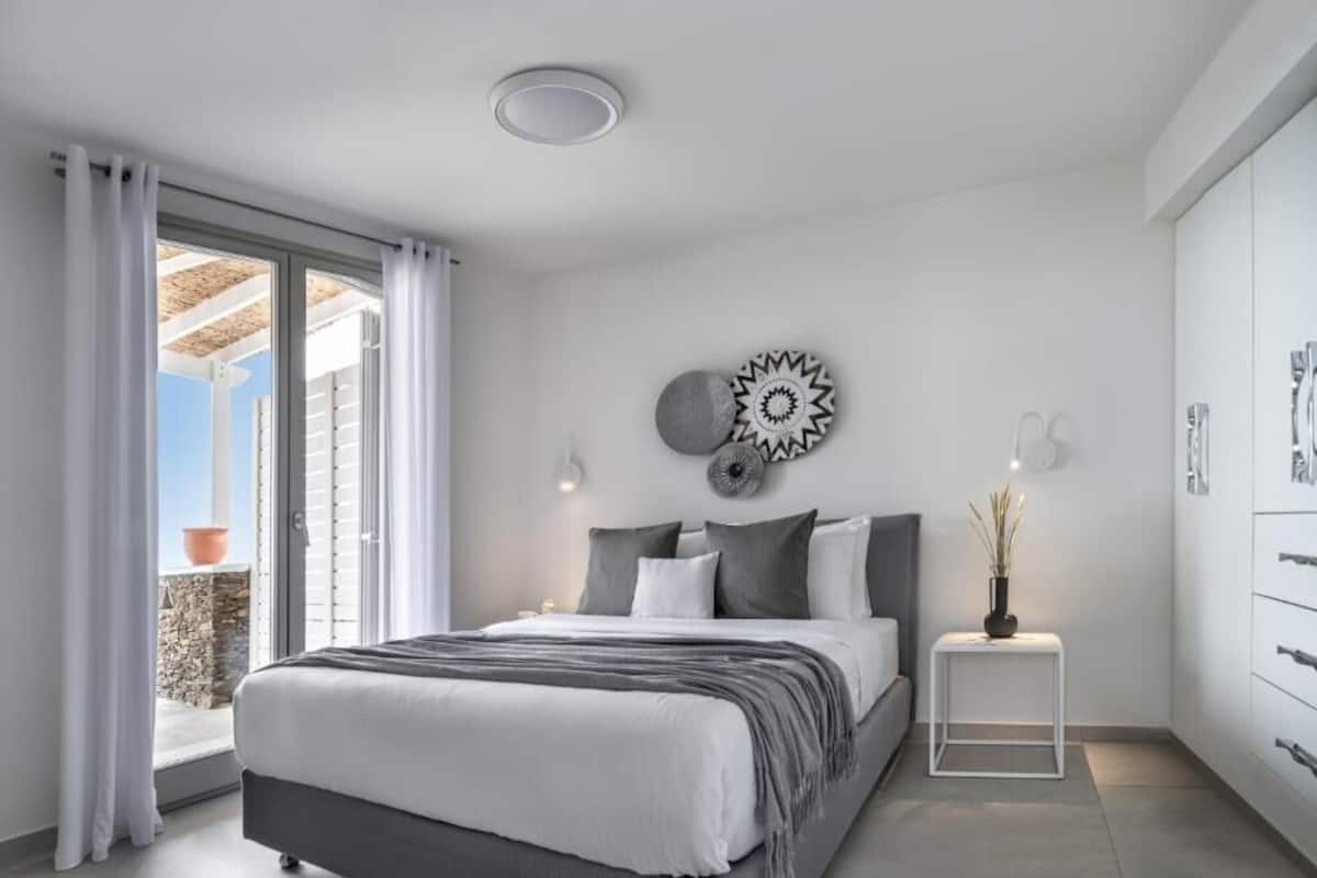A modern bedroom with a bed, a small table with a vase, and a view of the outside.