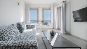 A modern living room with a sofa, a coffee table, and a TV mounted on the wall. There are two windows with curtains, a dining table with chairs, and a view of the sea. - NiMa Sifnos Residences (Sifnos)