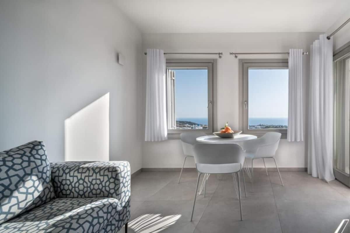 A modern living room with a patterned sofa, a round dining table with chairs, and large windows with curtains.