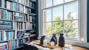 Quarto com uma grande estante repleta de livros, janela com vista para árvores e uma mesa com velas e vasos.