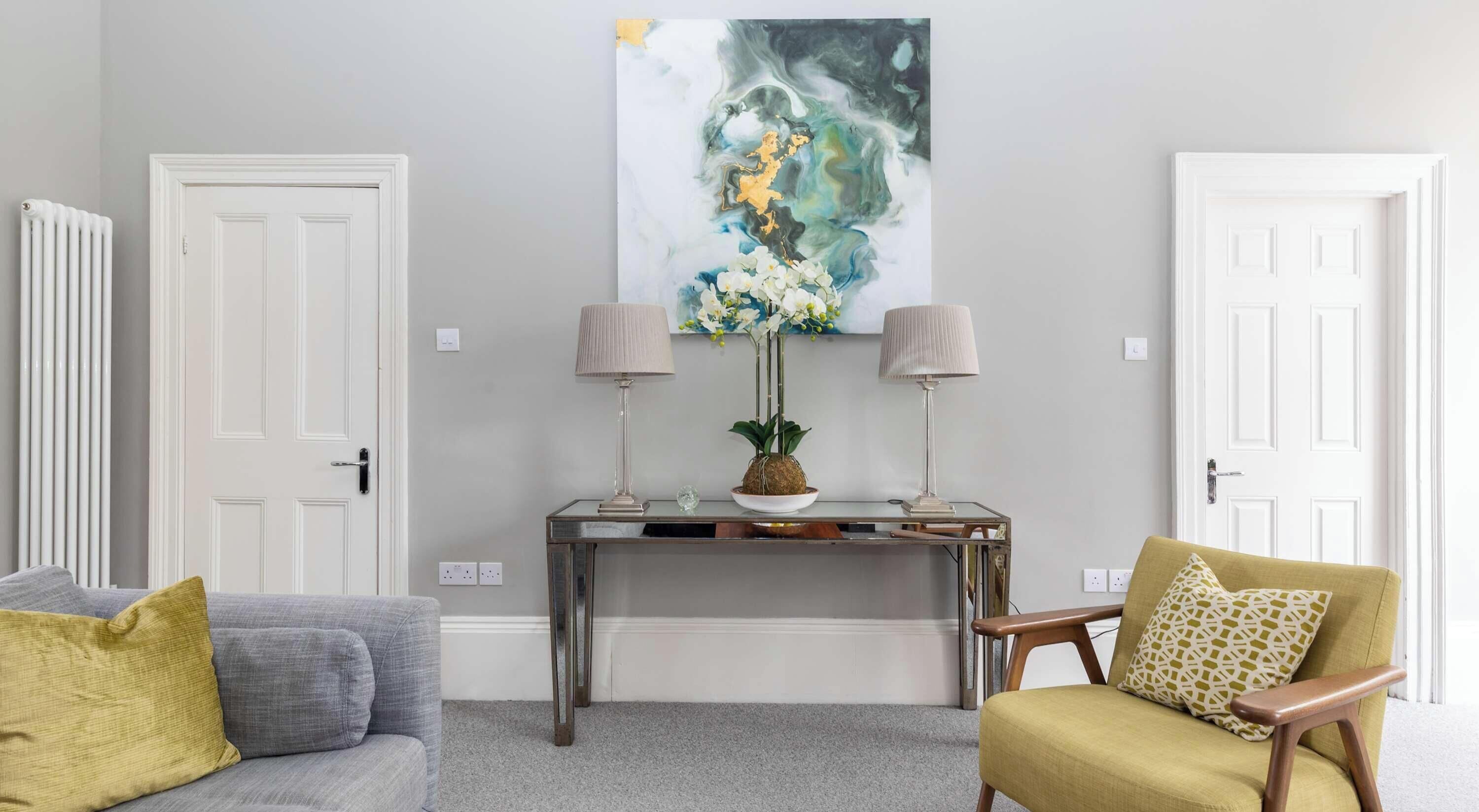 A living room with a grey sofa, a yellow armchair, a console table with two lamps, and a large abstract painting on the wall.
