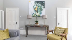 Ein Wohnzimmer mit grauem Sofa, einem gelben Sessel, einem Konsolentisch mit zwei Lampen und einem großen abstrakten Gemälde an der Wand.
