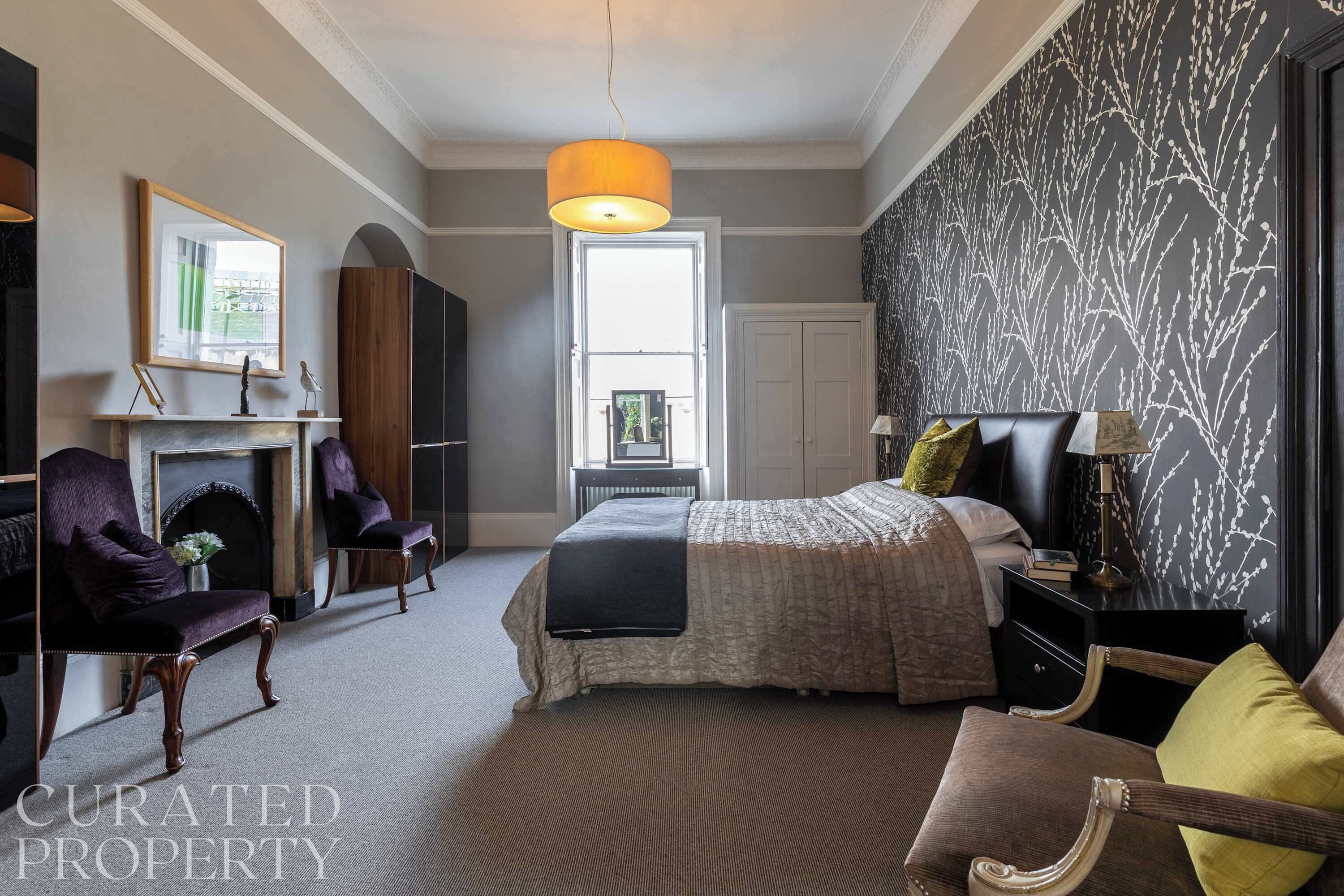 A bedroom with a bed, two armchairs, a nightstand, a lamp, a fireplace, and a large window.