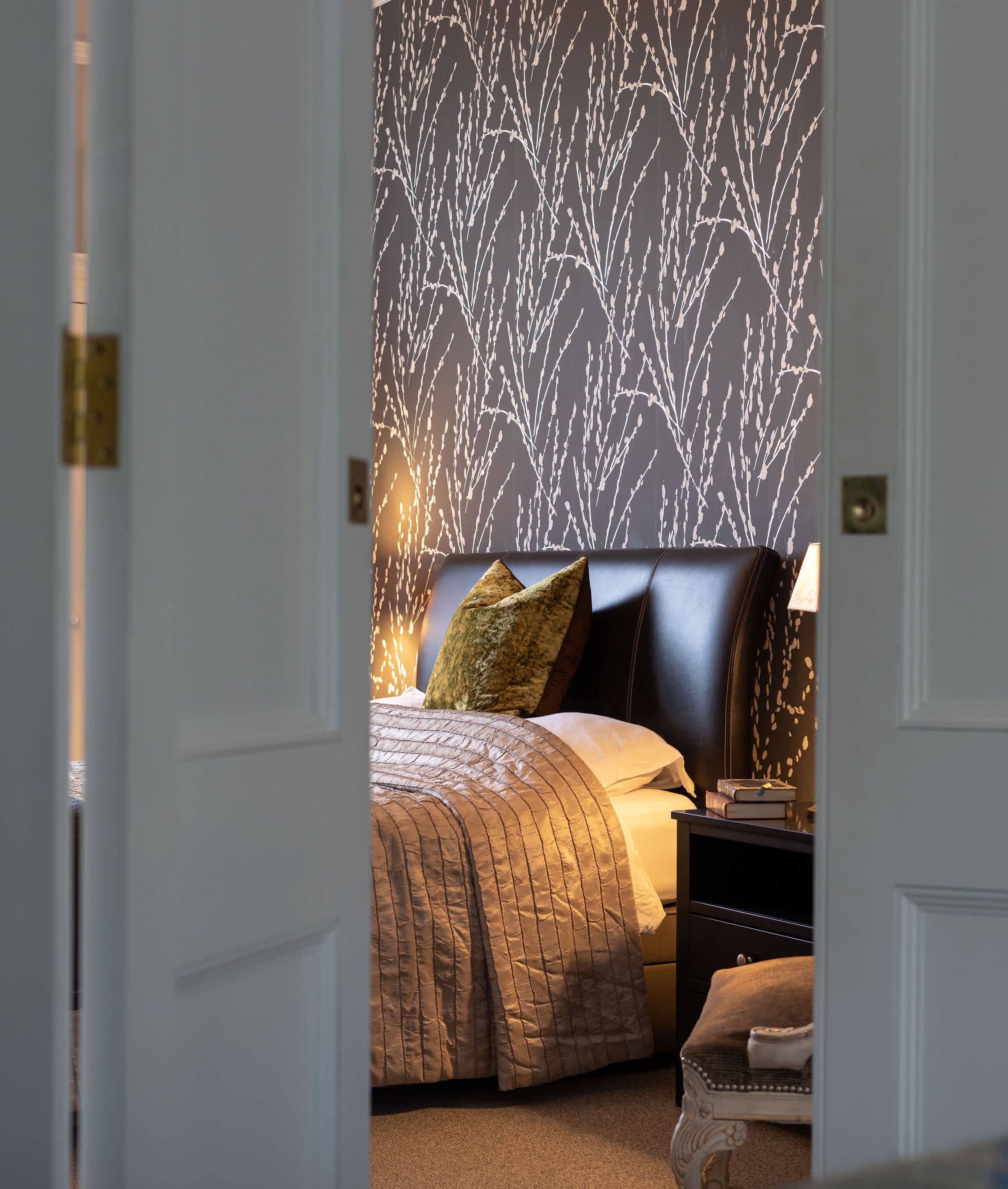 A bedroom with a patterned wallpaper, a bed with a headboard, pillows, and a bedside table with books.