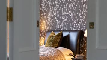 A bedroom with a patterned wallpaper, a bed with a headboard, pillows, and a bedside table with books.