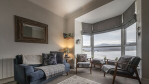 A cozy living room with a blue sofa, a wooden armchair, and a view of the sea through large windows. - Harbour View - Flat 2 - Tudor House (Barmouth)