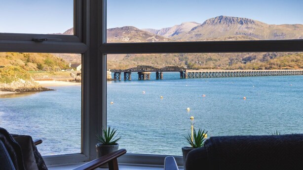 A view from a window overlooking a lake with a bridge and mountains in the background. - Harbour View - Flat 2 - Tudor House (Barmouth)