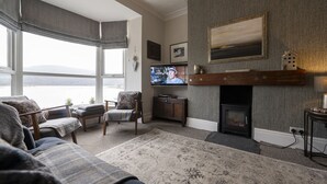 A living room with a fireplace, a television, and a large window overlooking a body of water. - Harbour View - Flat 2 - Tudor House (Barmouth)