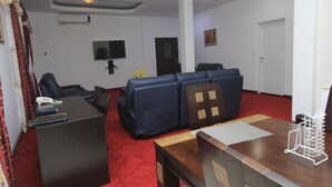 Un salon avec une moquette rouge, un bureau en bois foncé, un ensemble de canapés bleus, un téléviseur fixé au mur et une porte visible au fond.