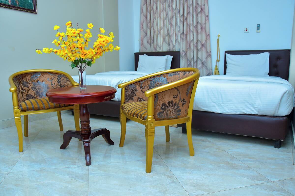 A hotel room with two yellow chairs, a small table, a bed with white bedding, and a vase with yellow flowers.
