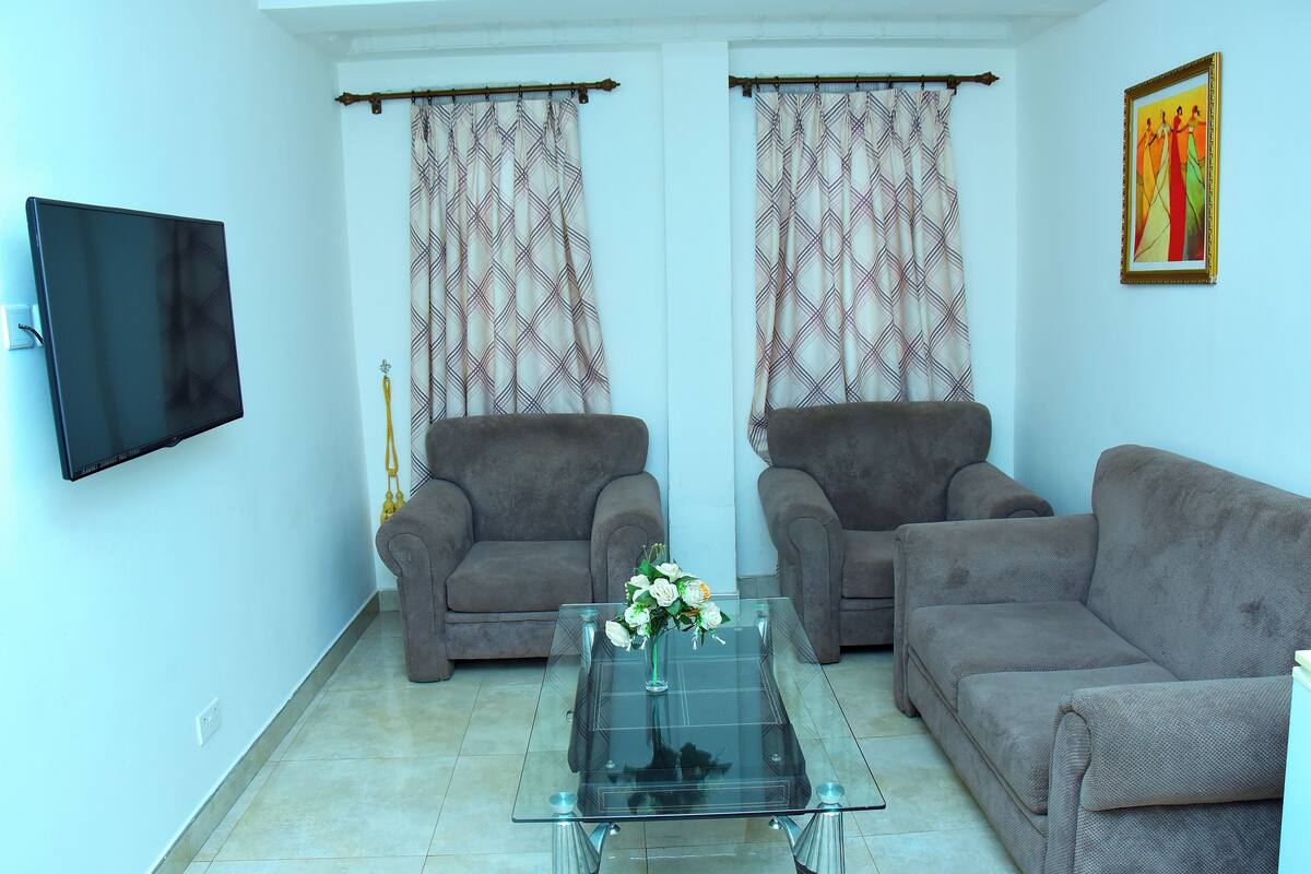 A living room with two grey sofas, a glass coffee table with a vase of flowers, and a flat-screen TV mounted on the wall.