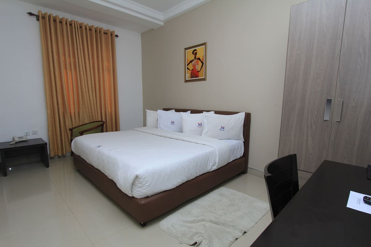 A hotel room with a bed, a desk, a chair, a wardrobe, and a painting on the wall.
