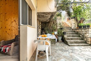 Un patio con suelo de piedra, una silla de plástico blanca, una mesa con fruta y una escalera que conduce a un muro de piedra.