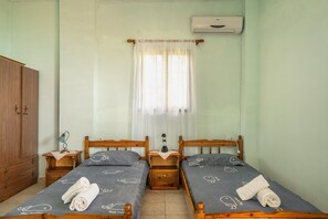 Une chambre avec deux lits simples, des têtes de lit en bois, une table de chevet avec une lampe, une armoire et une fenêtre avec un rideau léger.