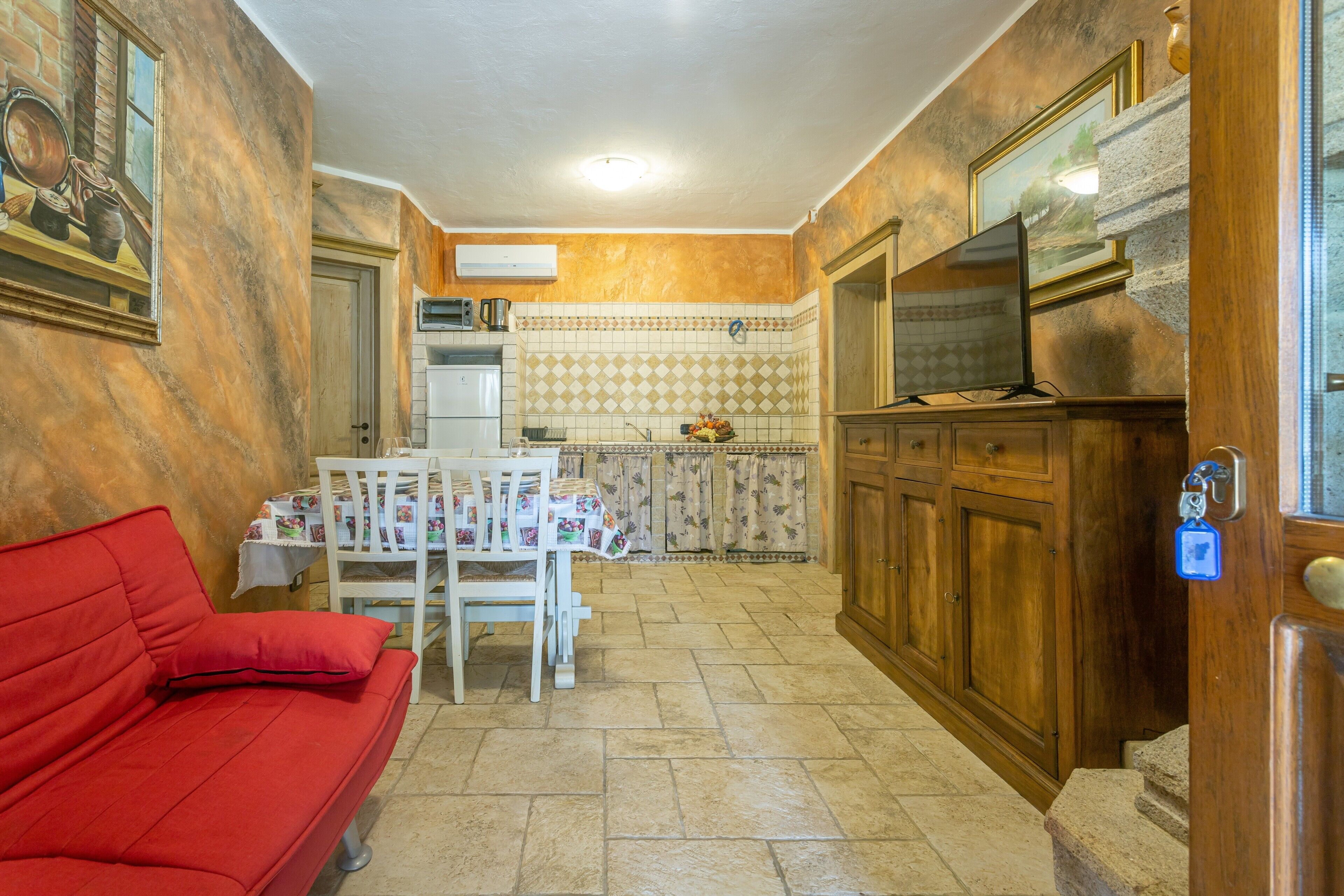 A compact kitchen with a dining area, a red sofa, and a TV mounted on the wall.