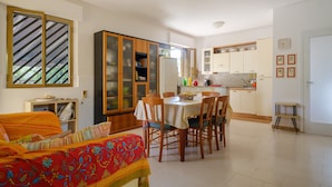 Un comedor con una mesa preparada para cuatro, una cocina con armarios blancos y un ventanal con persianas a rayas.