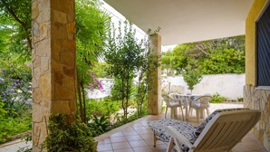 Un patio cubierto con pilares de piedra, una silla blanca, una mesita y abundante vegetación.