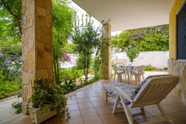 Un patio couvert avec des piliers en pierre, une chaise blanche, une petite table et une végétation luxuriante.