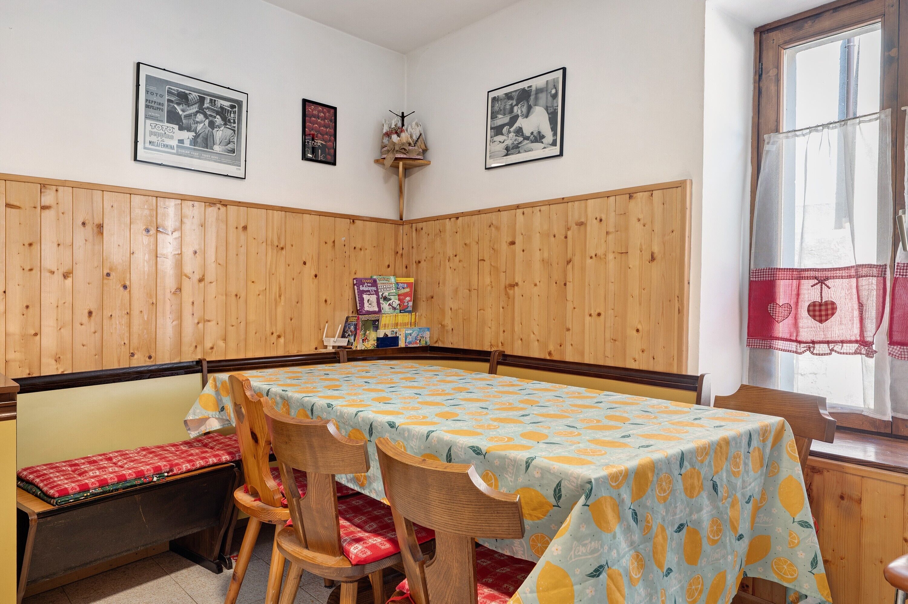 Ein Eckzimmer mit holzgetäfelten Wänden, einem Tisch mit Tischdecke mit Zitrusmuster, einer Bank mit roten Kissen und gerahmten Bildern an der Wand.
