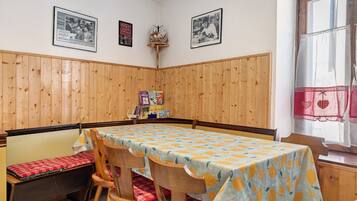 Ein Eckzimmer mit holzgetäfelten Wänden, einem Tisch mit Tischdecke mit Zitrusmuster, einer Bank mit roten Kissen und gerahmten Bildern an der Wand.