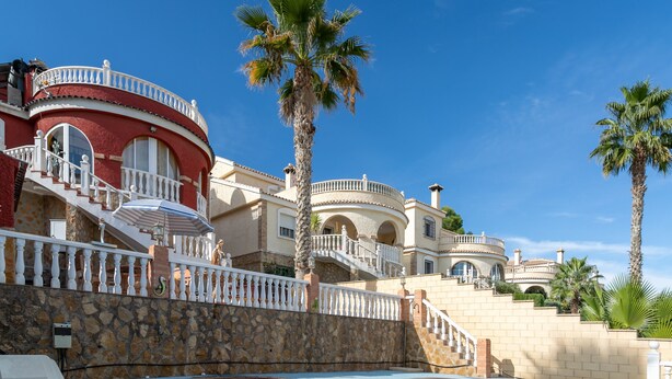 Une piscine bordée d’un mur en pierre et un escalier menant à un bâtiment à la façade rouge et aux balcons blancs.